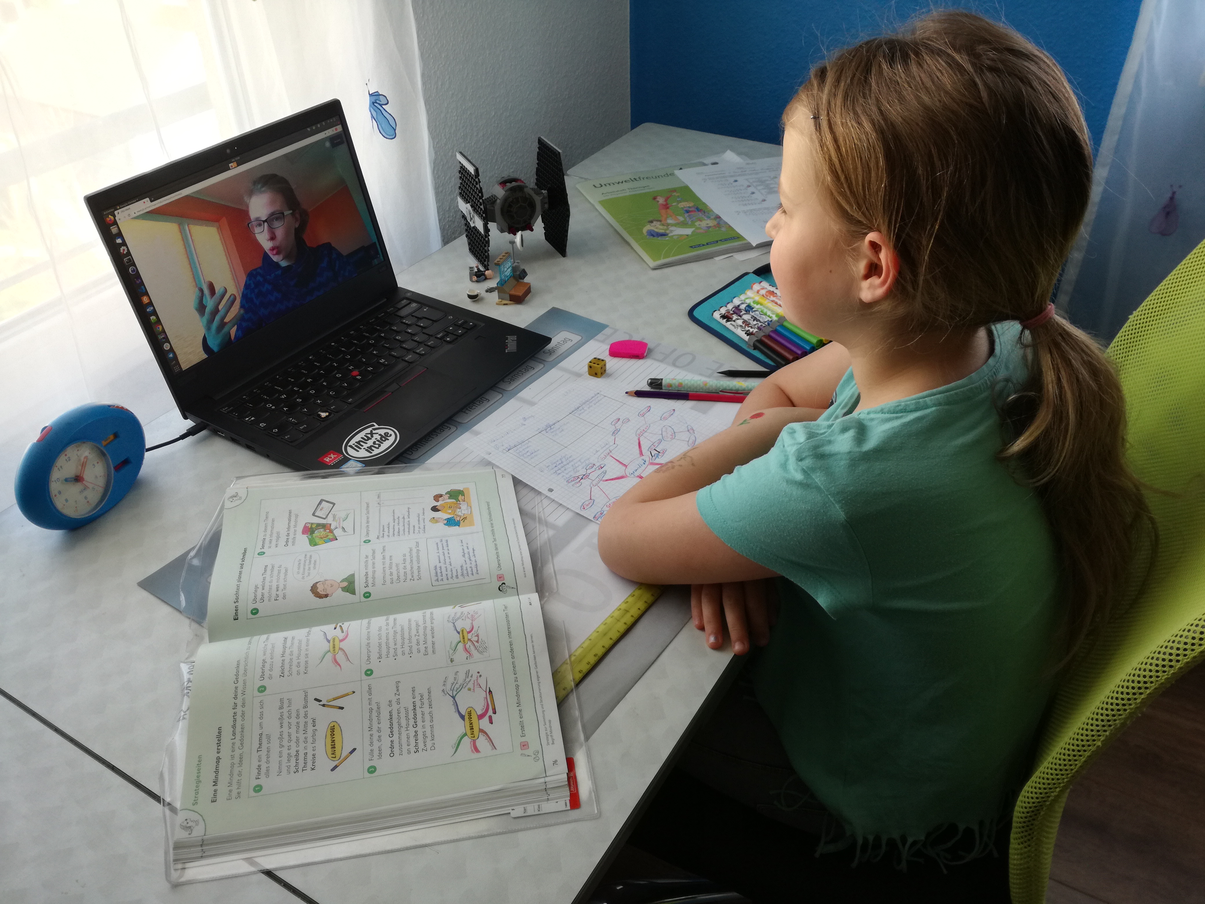 Mädchen mit aufgeschlagenen Schulheften bekommt am Laptop per Videokonferenz etwas von einer Studentin erklärt
