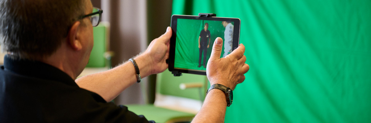 Eine Lehrkraft blickt durch die Kamera-App eines Tablets auf einen Green-Screen