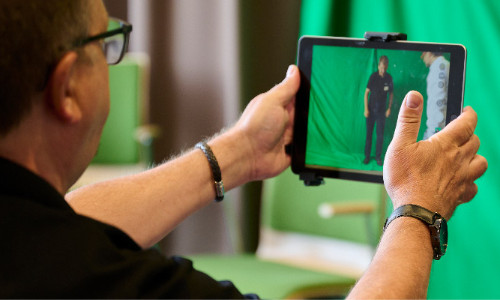 Eine Lehrkraft blickt durch die Kamera-App eines Tablets auf einen Green-Screen
