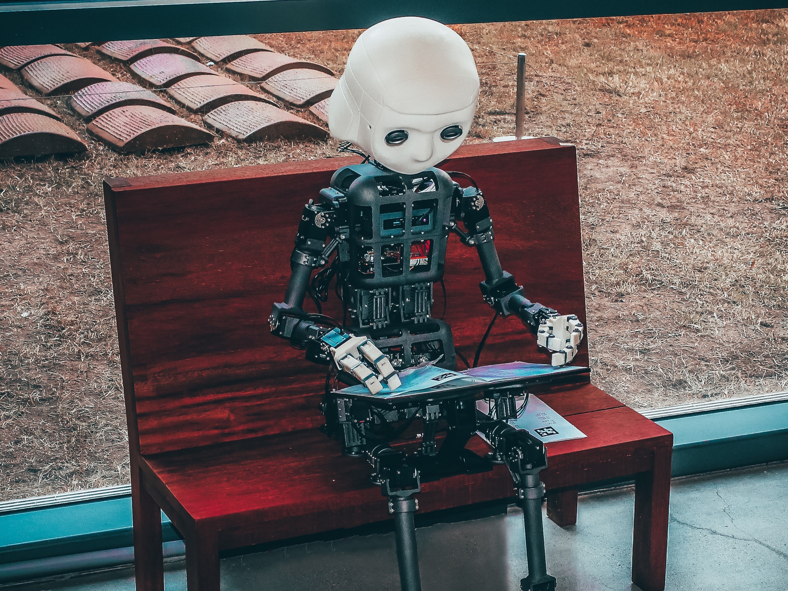 Roboter sitzt auf roter Bank und hält ein Tablet in den Händen. 