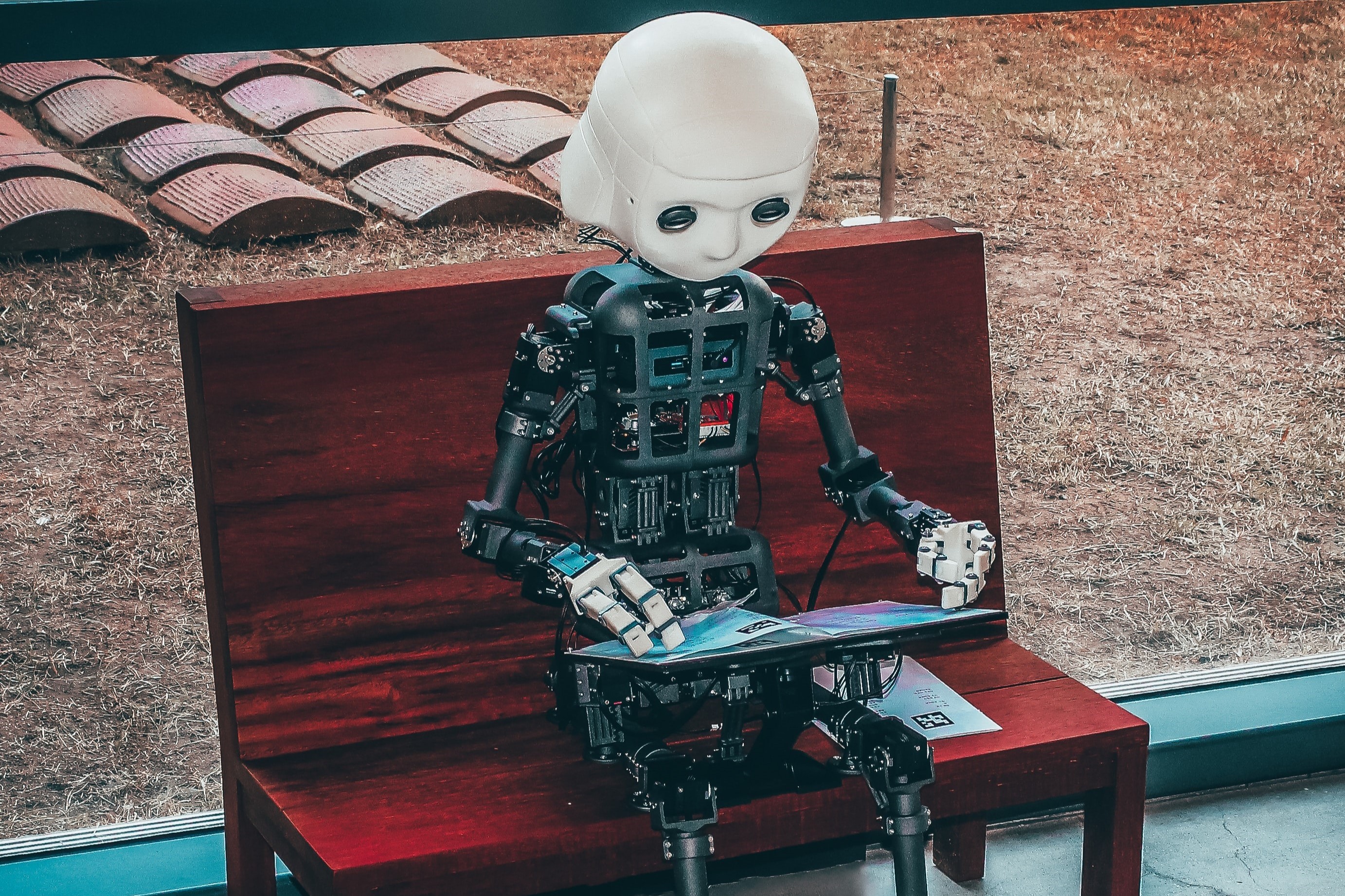 Ein Roboter sitzt auf einer roten Bank und hält ein Tablet in der Hand. 